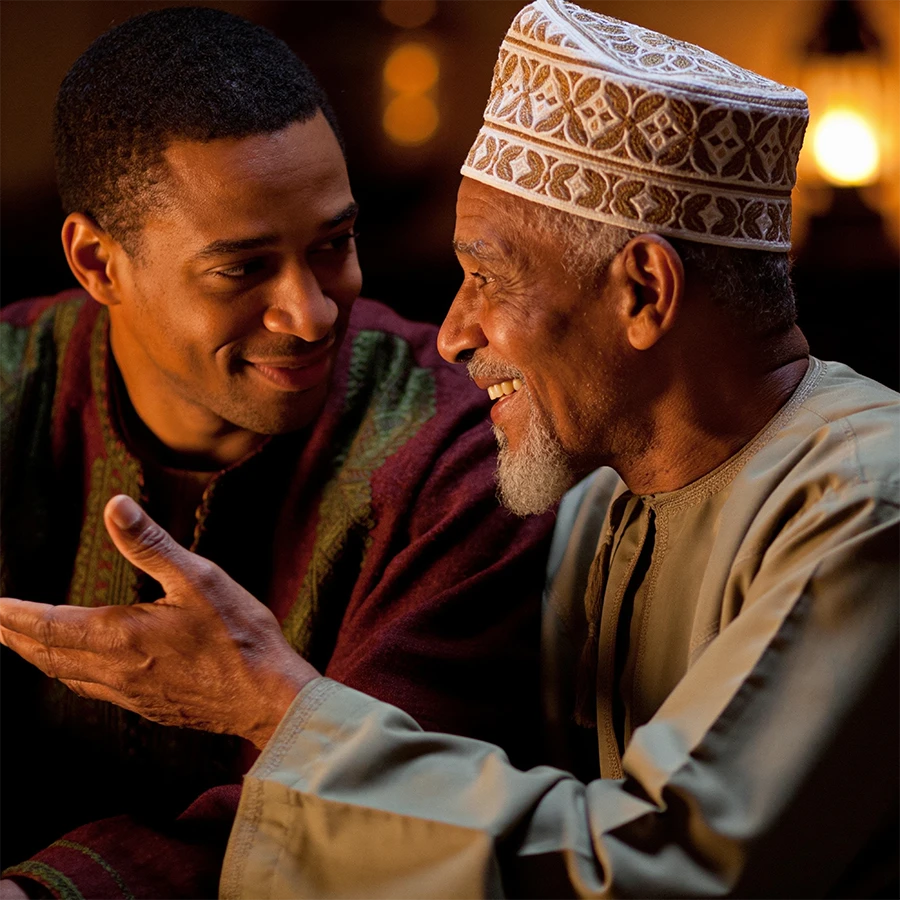 An elder shares a story with a younger man by lamplight